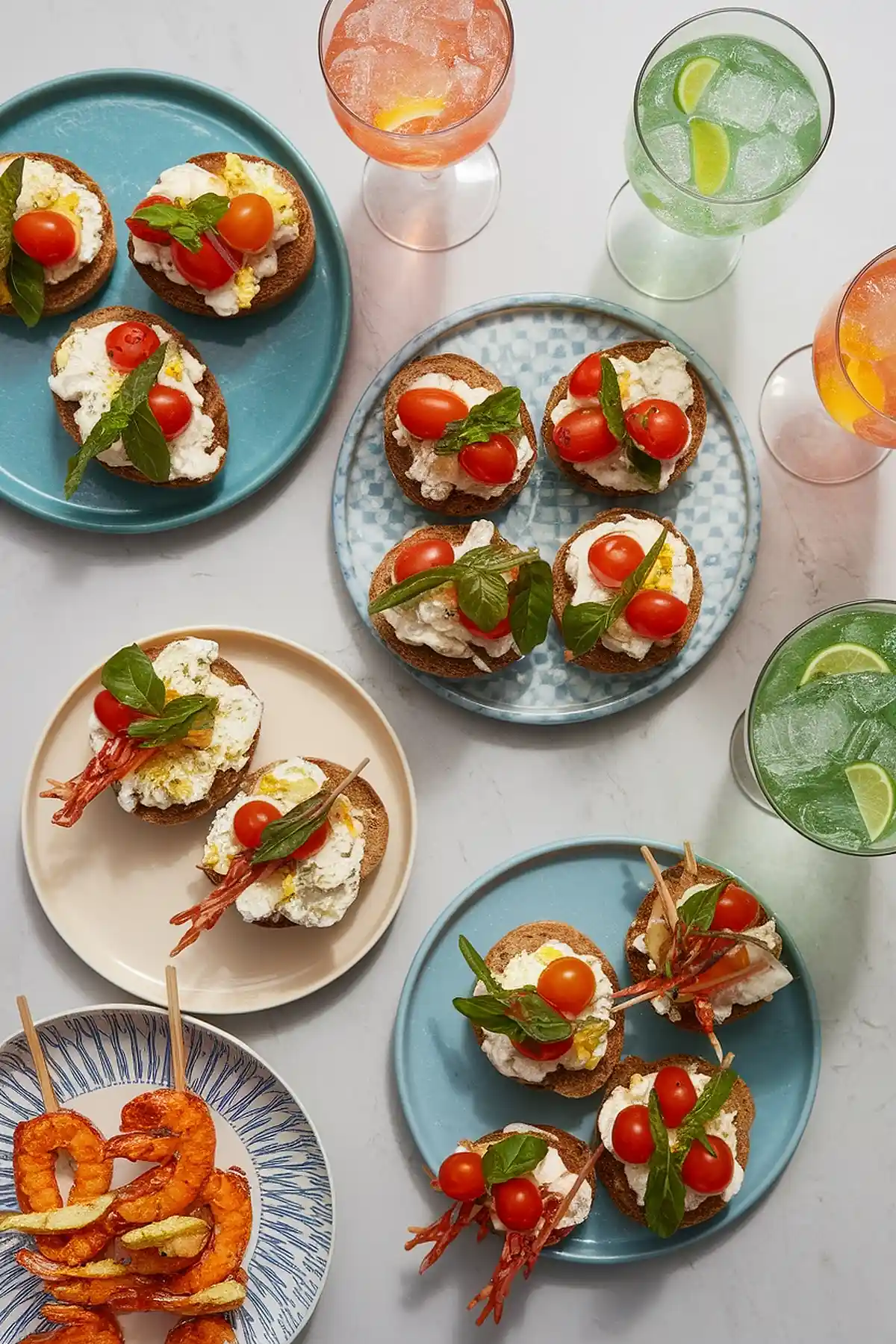 Assorted bruschetta with cherry tomatoes and shrimp beside colorful cocktails on a marble table.