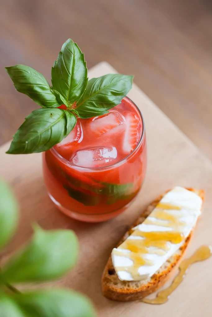 Refreshing tomato basil drink with ice, paired with honey-drizzled cheese crostini on a wooden board.