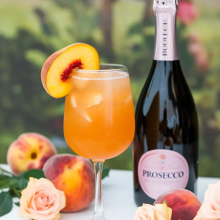 Peach Bellini cocktail with a slice of peach, Prosecco bottle, fresh peaches, and roses on a table.