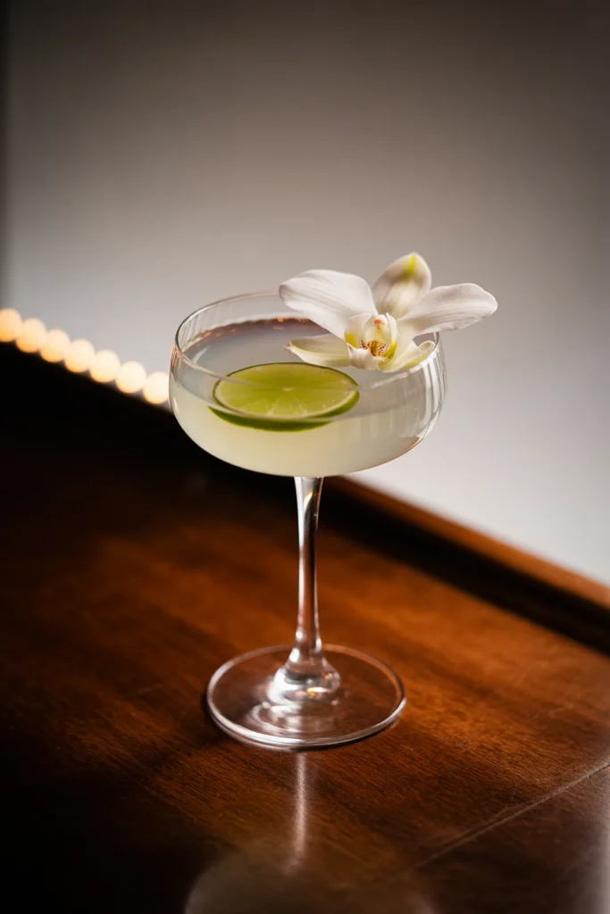 Elegant cocktail in a coupe glass with lime slice and orchid garnish on wooden surface.