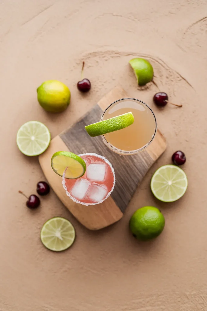 Refreshing lime cocktails with cherries and citrus slices on a wooden board, styled on a sandy surface.