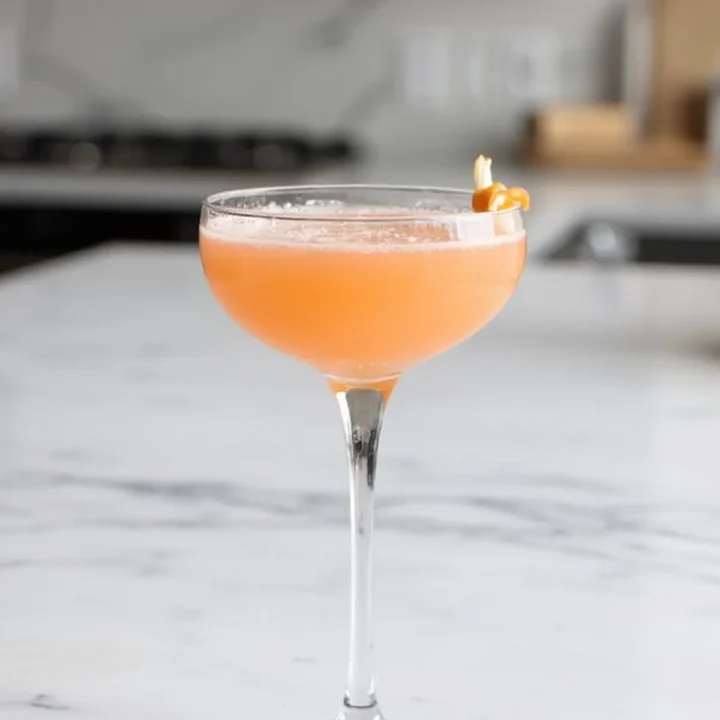 Elegant peach cocktail in a coupe glass on a marble countertop, garnished with citrus twist.